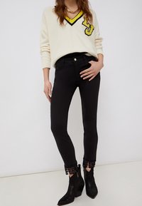 Žena oblečená v bílém svetr s výstřihem do V a žlutým a černým lemem, černých přiléhavých kalhotách se krajkovým lemem na kotnících a černých kotníkových botách.