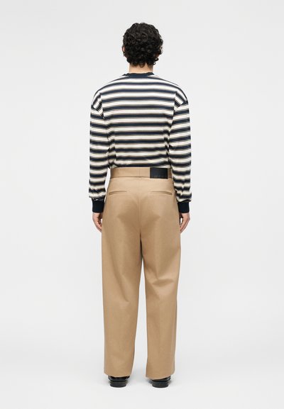 Άνδρας με σγουρά μαλλιά που κοιτάζει προς τα πίσω, φορώντας μια μπλε σκούρα και λευκή ριγέ μακρυμάνικη μπλούζα και μπεζ παντελόνι με ψηλή μέση και φαρδύ πόδι.