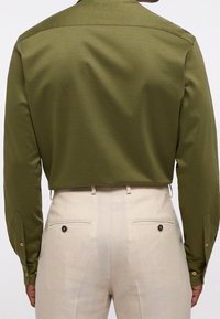 Camicia a maniche lunghe verde oliva, con texture e polsini a bottoni, abbinata a pantaloni beige con tasche posteriori e dettagli a bottoni.