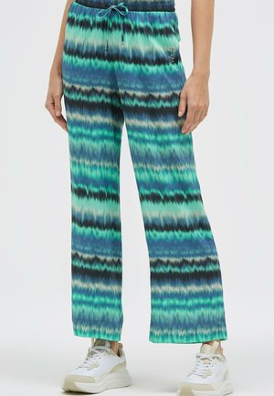 Frau trägt blau-grüne Batik-weit geschnittene Hosen mit Kordelzug in der Taille und weiße Sneakers, steht vor einfarbigem Hintergrund.