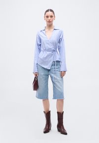 Μπλε και λευκό ριγέ πουκάμισο με μακριά μανίκια που δένει στο πλάι, συνδυασμένο με ανοιχτόχρωμες τζιν κολότσες. Το μοντέλο κρατά μια μπορντό τσάντα και φορά σκούρα καφέ μποτάκια καουμπόη.