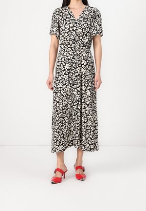 Kobieta ubrana w czarno-białą kwiatową sukienkę midi z krótkimi rękawami, dekoltem w serek oraz czerwone klapki na wysokim obcasie z metalicznymi noskami.