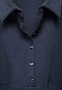 Devant de chemise bleu marine foncé avec col plié et quatre boutons noirs attachés le long de la patte sur un tissu texturé.