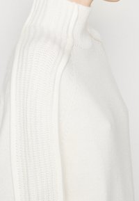 Vit stickad tröja med hög krage, ribbade detaljer längs axlarna och en mjuk textur. Närbild av sidovy.