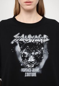 Svart grafisk t-skjorte med et panterdesign, hvit tekst som leser "SAUVAGE," og "VERSACE JEANS COUTURE" på et tydelig teksturert stoff.