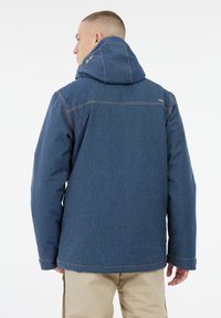 Mann mit kurzem Haar, der eine blaue Kapuzenjacke und beige Hosen trägt, mit dem Rücken zu einem weißen Hintergrund gewandt.