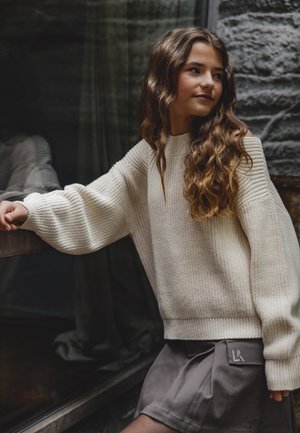 Cremfarbener, gestrickter Pullover mit weiten Ärmeln, kombiniert mit einem grauen, plissierten Rock, der ein dezentes Logo-Detail aufweist. Sanfte Texturen und lockere Passform.