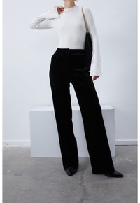 Biały prążkowany top z długim rękawem, ozdobiony guzikami przy dekolcie i mankietach, zestawiony z czarnymi welwetowymi spodniami o szerokich nogawkach oraz czarnymi szpilkami z ostrym noskiem.