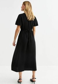 Vestido maxi negro con mangas cortas, cintura con cordón y una tela texturizada. Presenta una silueta relajada y fluida, con un dobladillo a la altura del tobillo.