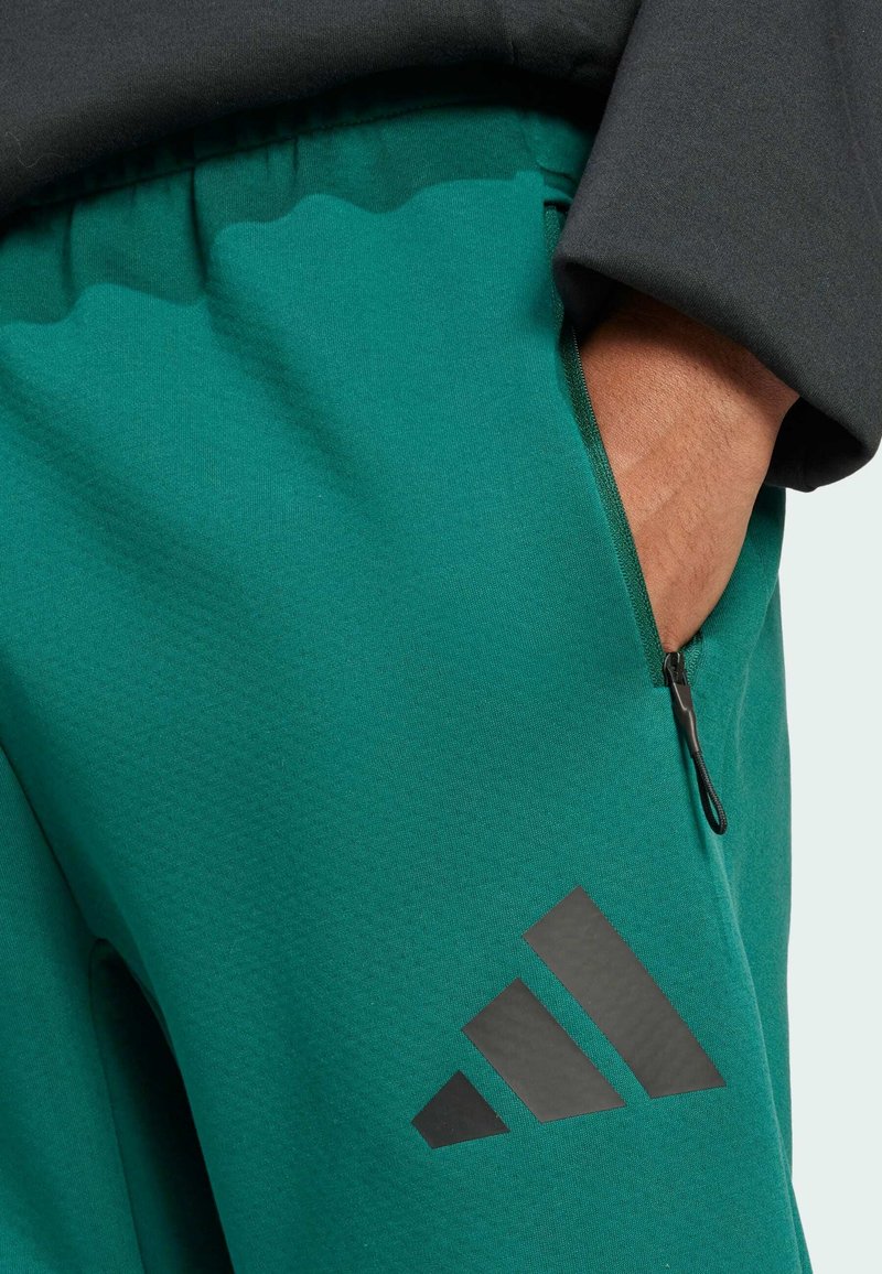 Zielone spodnie sportowe wykonane z gładkiego materiału o teksturowanym wykończeniu. Posiadają zapinaną na zamek kieszeń oraz duże czarne logo po boku.