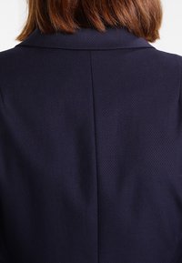 Veste bleu marine en tissu texturé, avec une fente simple au dos et un col classique, présentant des coutures lisses et sans éléments métalliques visibles.