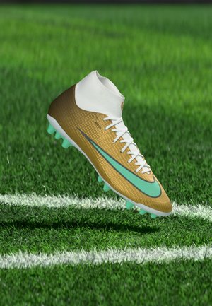 Gouden en witte voetbalschoen met een turquoise Nike-swoosh die boven een groen grasveld met witte grenslijnen zweeft.