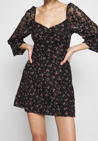 Mini-robe noire à fleurs avec corsage froncé, encolure carrée et manches bouffantes en tulle transparent ornées de détails en dentelle. Pose avec les mains sur les hanches.