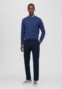 Suéter de punto azul marino sobre una camisa de collar a rayas azules y blancas, combinado con pantalones azul oscuro y zapatillas beige. Tela suave, corte casual.