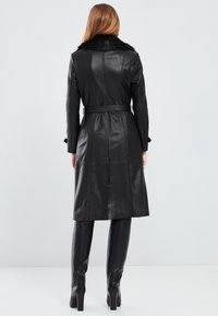 Cappotto lungo in pelle nera con colletto in pelliccia sintetica, vita con cintura e cuciture verticali; abbinato a stivali neri con tacco alto.