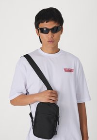 Black crossbody bag with a smooth texture, zippers, and an adjustable strap, worn over a white t-shirt with a red graphic.