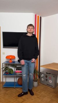 Νέος άνδρας με μαύρο φούτερ με κουκούλα και σκισμένα τζιν στέκεται σε ένα δωμάτιο με τηλεόραση, πορτοκαλί λαμπατέρ, φυτά σε γλάστρες, μπάλα μπάσκετ και βαλίτσα με αυτοκόλλητα.