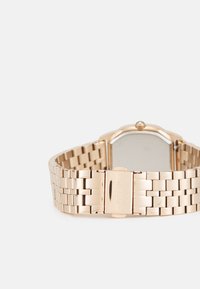Montre-bracelet en métal doré avec un bracelet à maillons, boîtier rectangulaire et surface lisse. Le fermoir présente le nom de la marque gravé.