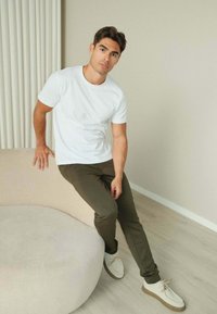 Hvid T-shirt med korte ærmer, olivengrønne bukser og hvide sko med brune såler. Sidder på et buet beige sofa med en neutral baggrund.