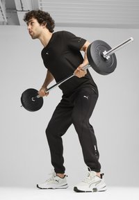 Camiseta atlética negra con logo, pantalones de chándal negros y zapatillas blancas. Sosteniendo una barra con pesas negras en una postura de entrenamiento.