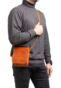 Borsa a tracolla in pelle marrone con forma strutturata, tracolla regolabile e dettagli di cucitura visibili, indossata da una persona in un maglione grigio.