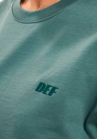 Grünes Baumwoll-T-Shirt mit Rundhalsausschnitt, das auf der linken Seite ein strukturiertes "DEF"-Logo in dunklem Grün aufweist, gefertigt aus einem glatten, atmungsaktiven Material.