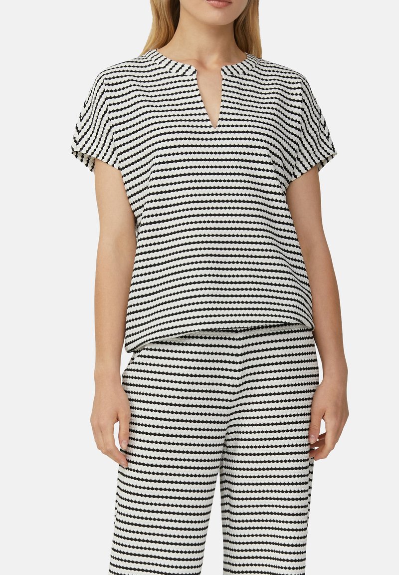 Bluzka z krótkim rękawem w czarno-białym materiale z geometrycznym wzorem. Posiada dekolt w serek i luźny krój, zestawiona z pasującymi spodniami.