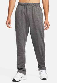 Homme portant un pantalon de survêtement gris Nike et des baskets blanches Nike, debout les bras détendus le long du corps sur un fond blanc.