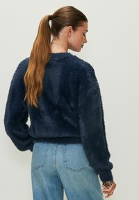 Maglione fuzzy blu marino con maniche lunghe e bordo a costine; indossato con jeans azzurri chiari. Tessuto morbido e strutturato con una vestibilità comoda.