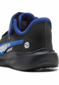 Chaussure de sport noire avec une bande accent bleu, dotée d'un col rembourré, d'une bride en Velcro et d'un détail de logo rond près du talon.