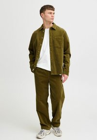 Olive green corduroy shirt with a button front and chest pocket, paired with matching pants. White t-shirt underneath and neutral sneakers.
