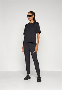 Svart sportoutfit bestående av en lös t-shirt och tajta byxor med varumärkeslogotyp. Accessoarer inkluderar mörka solglasögon och svarta skor.