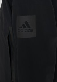 Tissu noir avec un patch du logo Adidas réfléchissant foncé cousu sur la manche ou le panneau latéral du vêtement.
