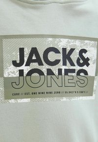 Felpa grigia chiara con una grafica prominente in verde scuro e bianco. Il testo include "JACK & JONES" e le coordinate. Texture in cotone morbido.