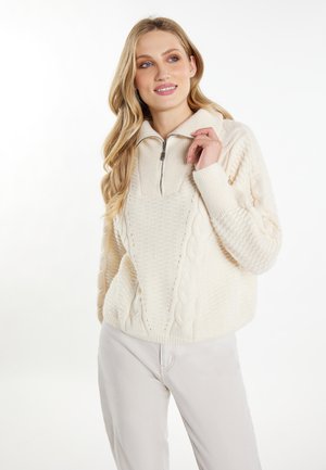 Donna bionda che indossa un maglione crema lavorato a trecce con cerniera a un quarto e pantaloni chiari, sorridente e con lo sguardo leggermente di lato.
