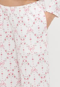 Biely textil s kvetinovým vzorom, ktorý zobrazuje ružové ruže, sivé viniče a ružové mašle. Obsahuje elastický pás a bočnú kapsu.