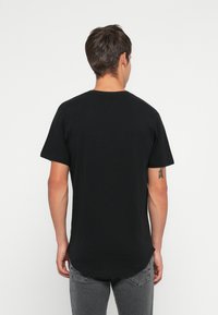 Hombre con cabello corto y oscuro que lleva una camiseta negra lisa y jeans grises, de espaldas, mostrando un tatuaje de flecha en el brazo izquierdo.