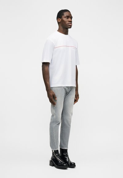 Άνδρας που φοράει λευκό πουκάμισο με κοντά μανίκια και κόκκινη γραμμή, ανοιχτό γκρι τζιν και γυαλιστερά μαύρα μποτάκια, στέκεται μπροστά σε απλό λευκό φόντο.