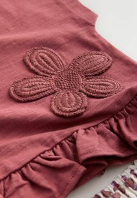 Close-up af pink stof med broderet femblomstret blomst og rynket flæsedetalje på beklædningsgenstand.