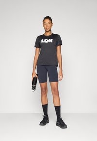 T-shirt de sport noire avec le texte "LDN", associée à un short sombre et des chaussures de sport noires. Le mannequin tient une bouteille d'eau.