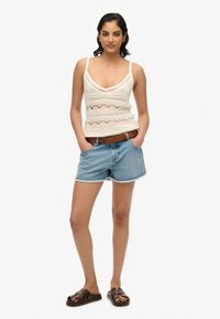 Top de tirantes de ganchillo en color crema con un escote en V profundo, combinado con pantalones cortos de mezclilla azul claro con dobladillos deshilachados, y sandalias tipo slide en color marrón.