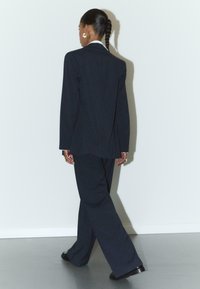 Navy-blaues Nadelstreifenanzug mit einem maßgeschneiderten Sakko und weit geschnittenen Hosen. Glattes Material, strukturierter Schnitt und klassisches Detail mit weißem Kragen.