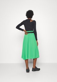 Haut noir à manches longues avec un détail découpé, associé à une jupe verte plissée arrivant au genou et des chaussures de sport noires.
