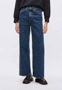 Modré široké džínsové nohavice s vysokým pásom, vybavené piatimi vreckami, kontrastným prešívaním a neupravenými lemami. Nosené s čiernymi platformovými topánkami.