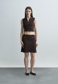 Štíhla žena stojí oblečená v bezrukávovom hnedom pletenom krátkom topánku a zodpovedajúcej sukni s vlnovitými vzormi a čiernych balerínach na sivom podlahe.