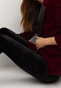 Bordeauxrode fluwelen blazer met een zwarte satijnen revers, gecombineerd met zwarte fluwelen leggings. Soepele textuur en aansluitend ontwerp, zittend tegen een neutrale achtergrond.