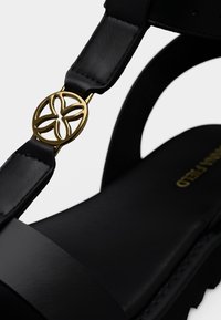 Μαύρο δερμάτινο σανδάλι με χρυσή κυκλική λεπτομέρεια που παρουσιάζει floral σχέδιο στο λουρί. Ομαλή υφή με επίπεδη, μαλακή σόλα.