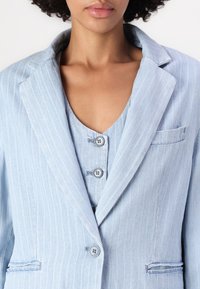 Blazer a righe sottili blu chiaro con colletto a scollatura, tasca singola e due bottoni, indossato sopra un top senza maniche coordinato.