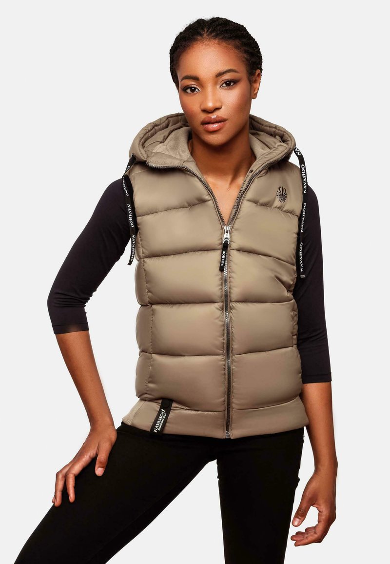 Beige Steppweste mit Reißverschluss und Kapuze, die ein elegantes Design, vertikale Nähte und markierte Kordeln aufweist. Getragen über einem schwarzen Oberteil.