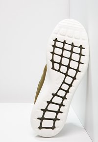 Nike-sko med en vit gummisula som har ett strukturerat greppmönster. Mörk olivgrön ovandel synlig längs kanten.